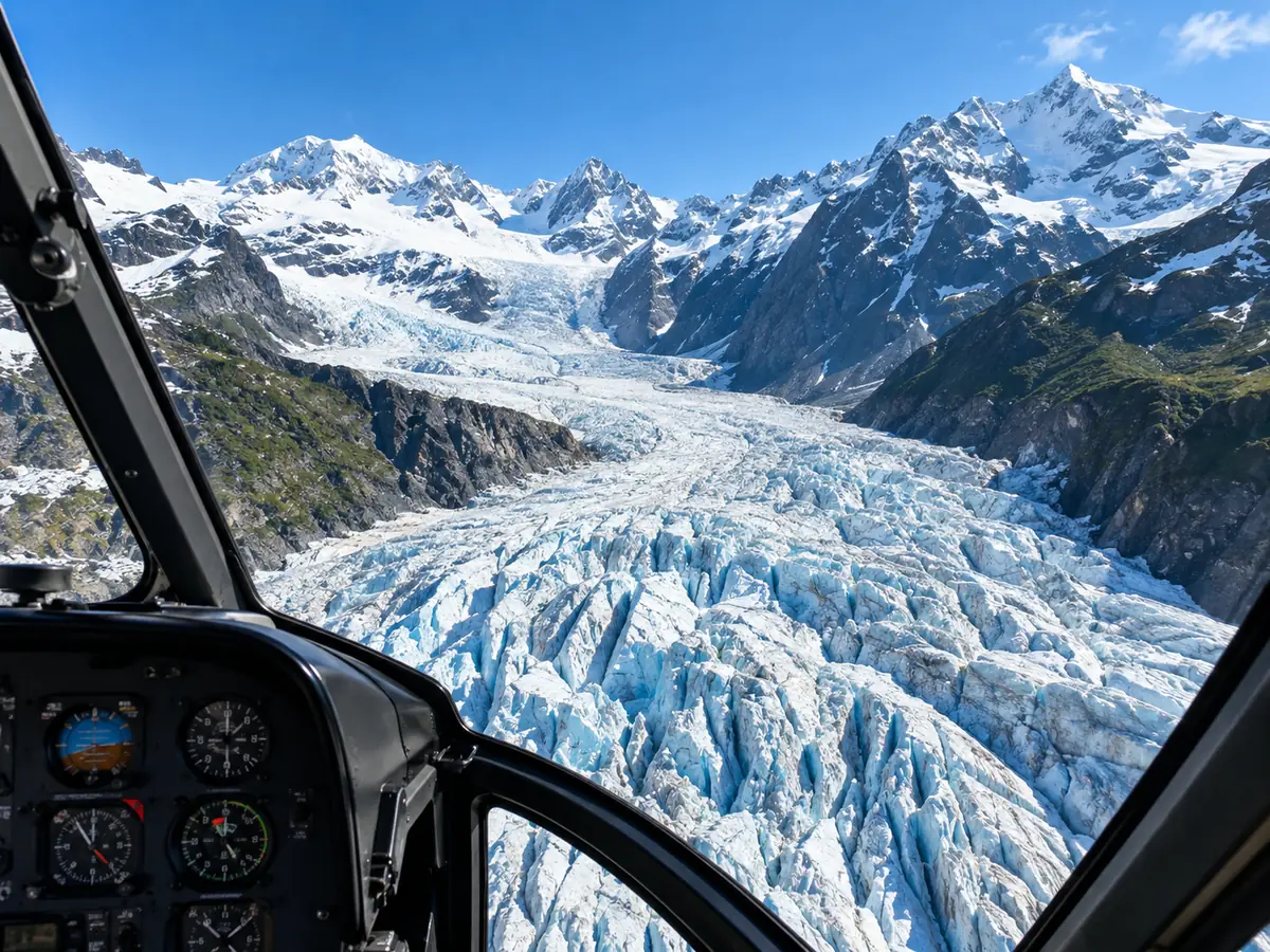 Blick aus einem Hubschrauber-Cockpit über einen zerklüfteten Gletscher und verschneite Gipfel unter blauem Himmel