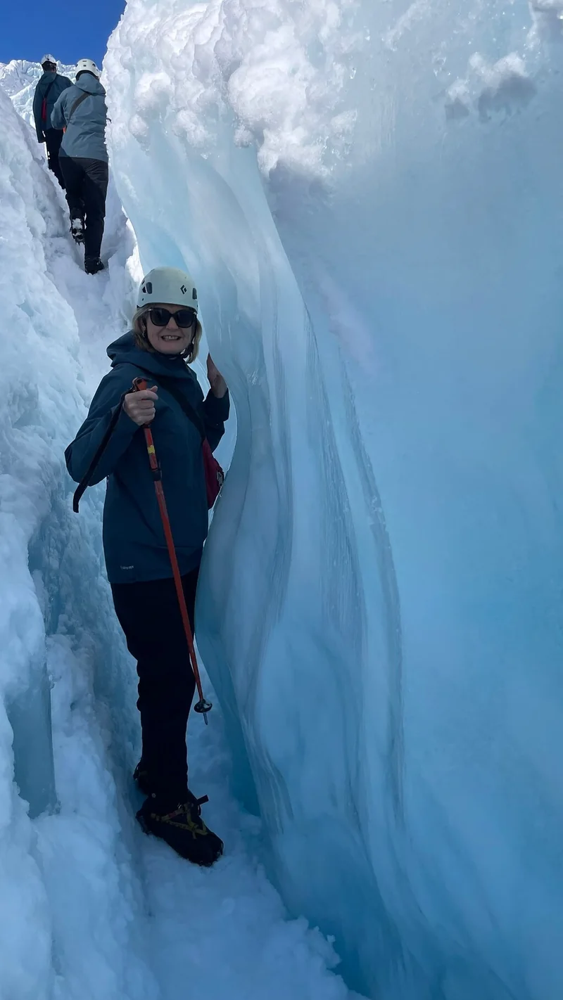 A hiker in a helmet, sunglasses and blue waterproof jacket standing in a narrow ice crevasse holding a trekking pole