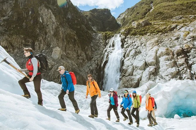 Eine Reihe Wanderer in Steigeisen geht auf dem Fox Glacier neben einem Wasserfall, der von einer Felswand stürzt